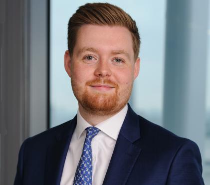 man with ginger hair and beard, wearing navy suit, white shirt, blue tie smiling